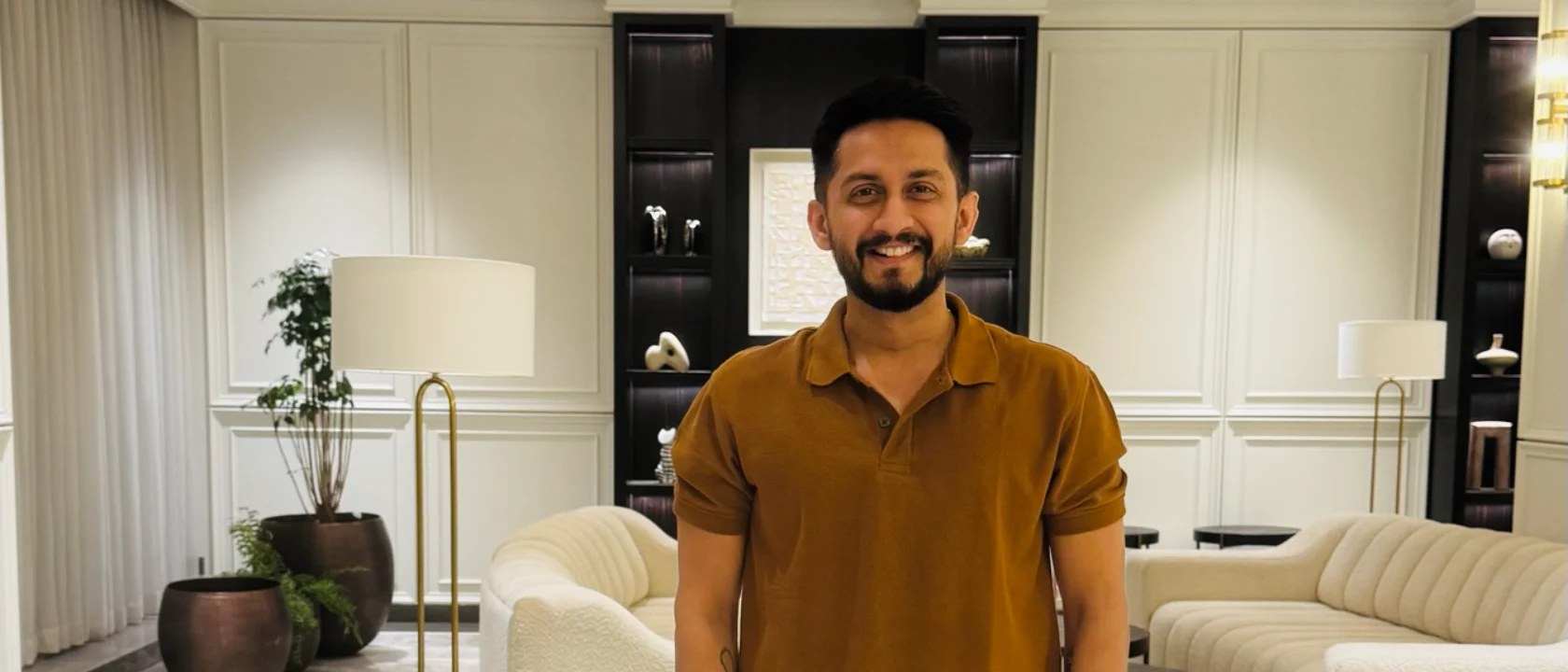 Digital Pratik, a man in a brown polo and cream trousers stands centered in a hotel lobby lounge, smiling broadly at the camera