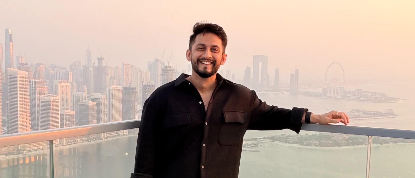 Digital Pratik smiling on the viewing deck at The View at The Palm, the Dubai Marina skyline and the sea behind him at sunset