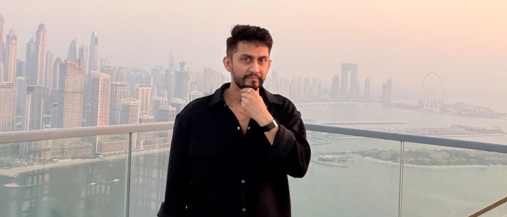 Digital Pratik, the man stands upright facing the camera, hand near his chin, with a serious look against the skyline backdrop. taken at sunset on the viewing deck at the top of the Palm tower (The View at The Palm), with the Dubai Marina skyline and the sea behind him