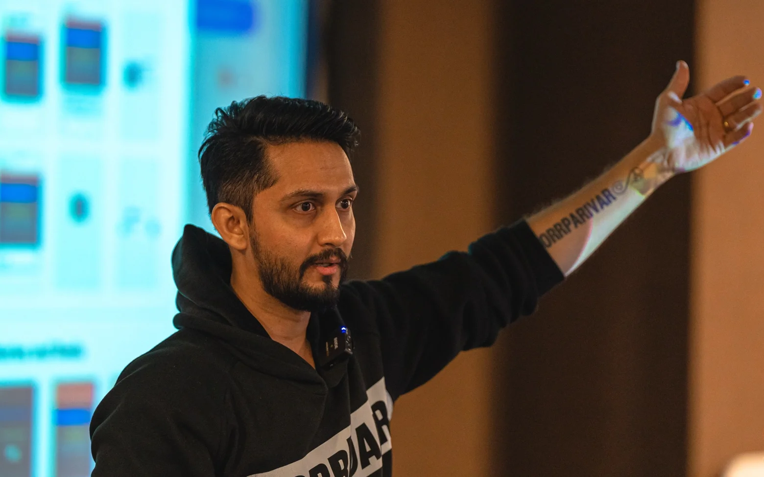 Digital Pratik teaching at a workshop in a black JorrDaar hoodie, an arm stretched out toward a screen behind him