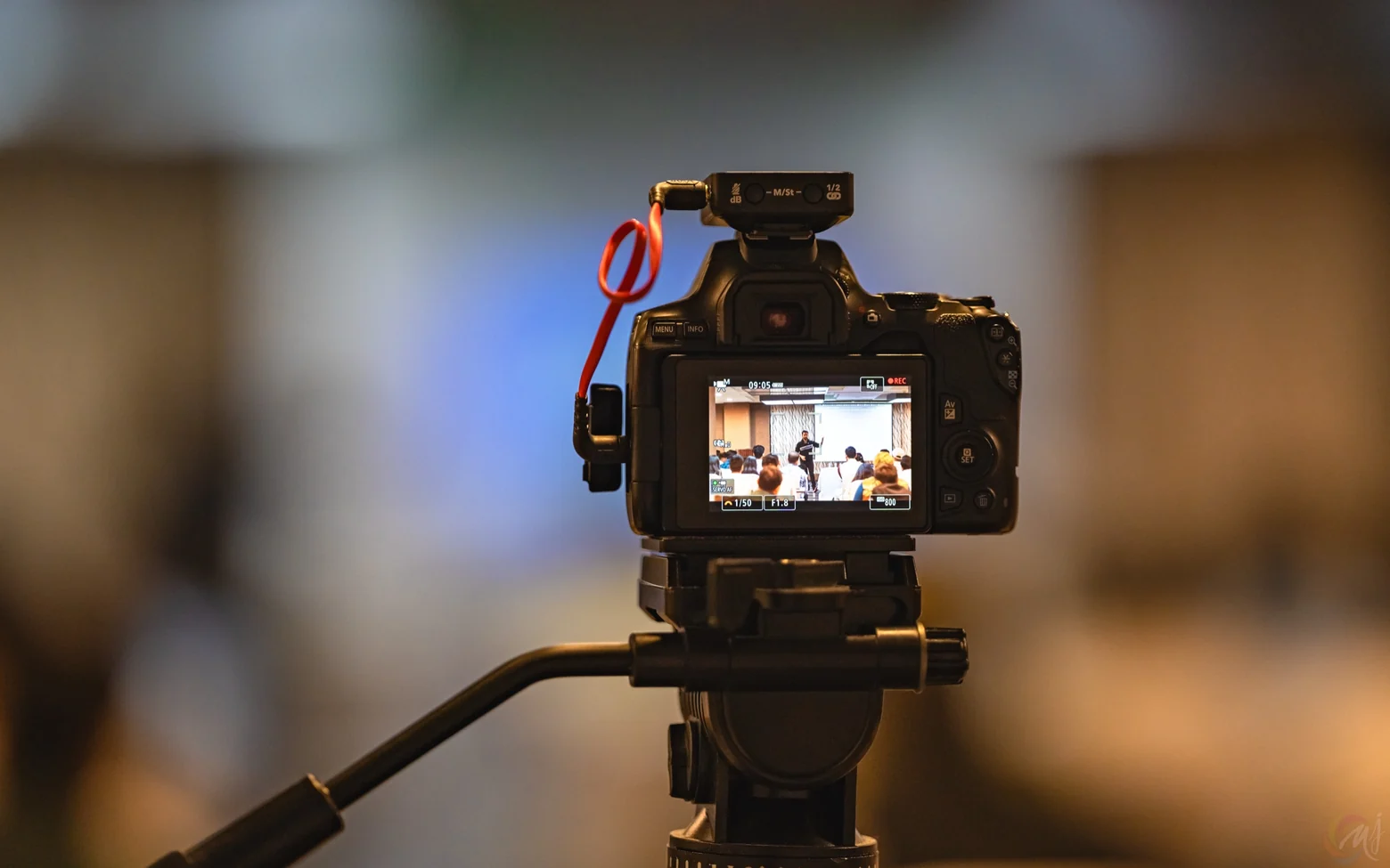 A camera on a tripod filming Digital Pratik's March 2024 workshop, the room and the stage small on its screen