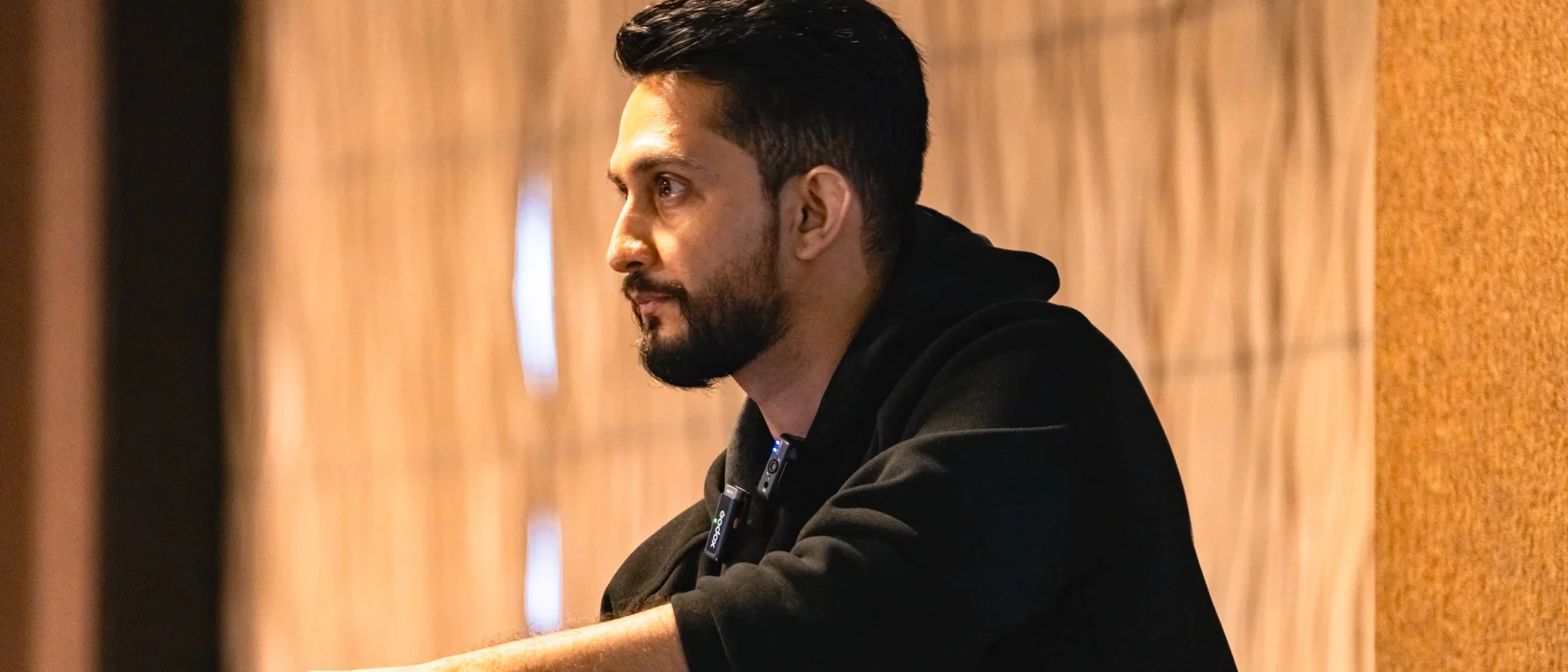 Digital Pratik in a black hoodie, looking across the room and listening, a warm wooden wall behind him