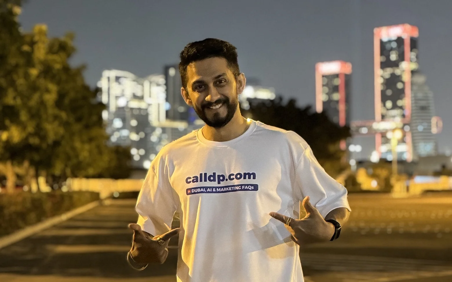 Digital Pratik smiling at night in a white calldp.com t-shirt, lit city towers behind him