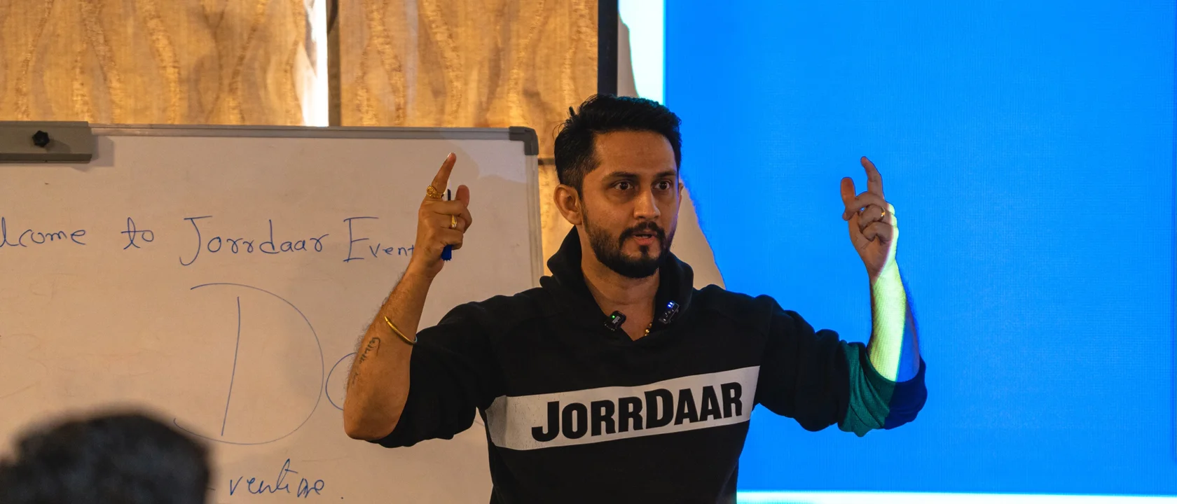 Digital Pratik teaching a room in a black JorrDaar hoodie, both hands up, a whiteboard and a blue screen behind him