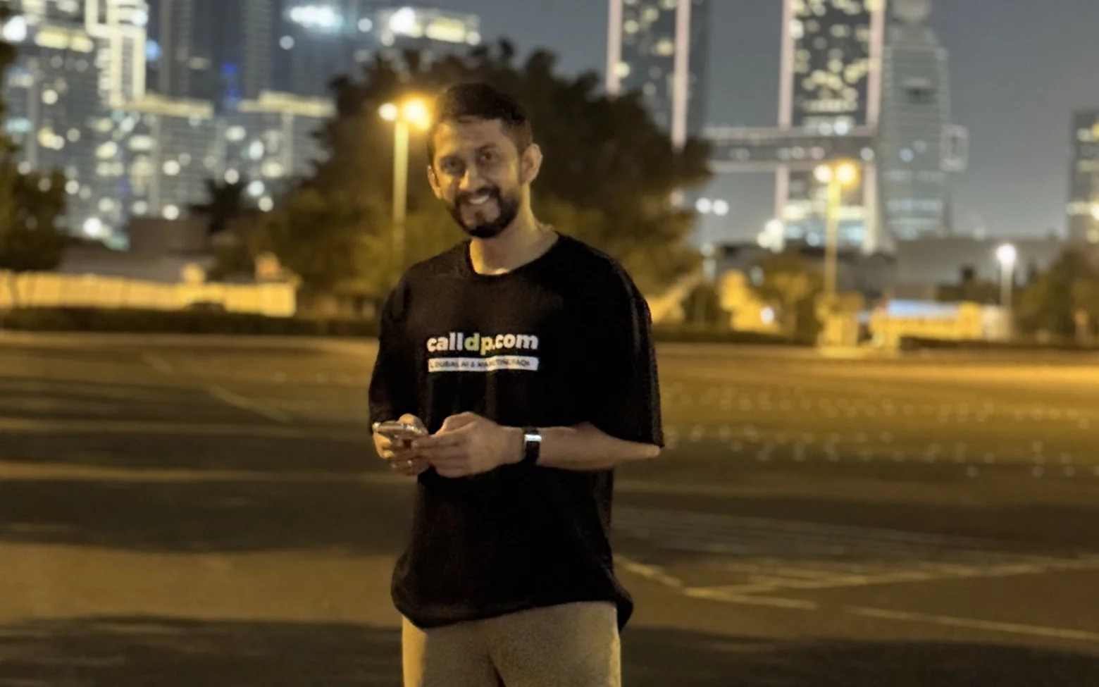 Digital Pratik smiling at night in an empty car park, holding his phone, city towers behind him