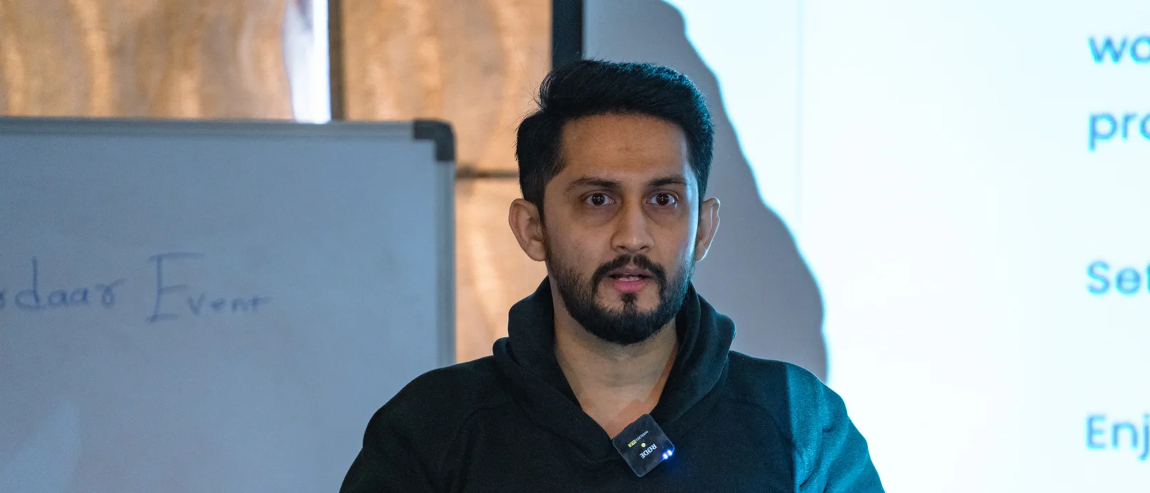 Digital Pratik speaking at a workshop in a black hoodie with a mic on his collar, a whiteboard and a projected slide behind him