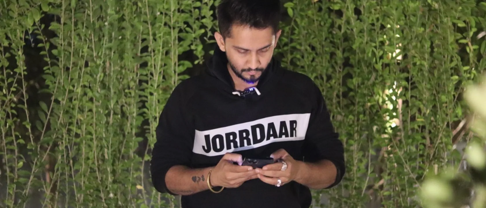 Digital Pratik typing on his phone in a black JorrDaar hoodie, green leaves behind him