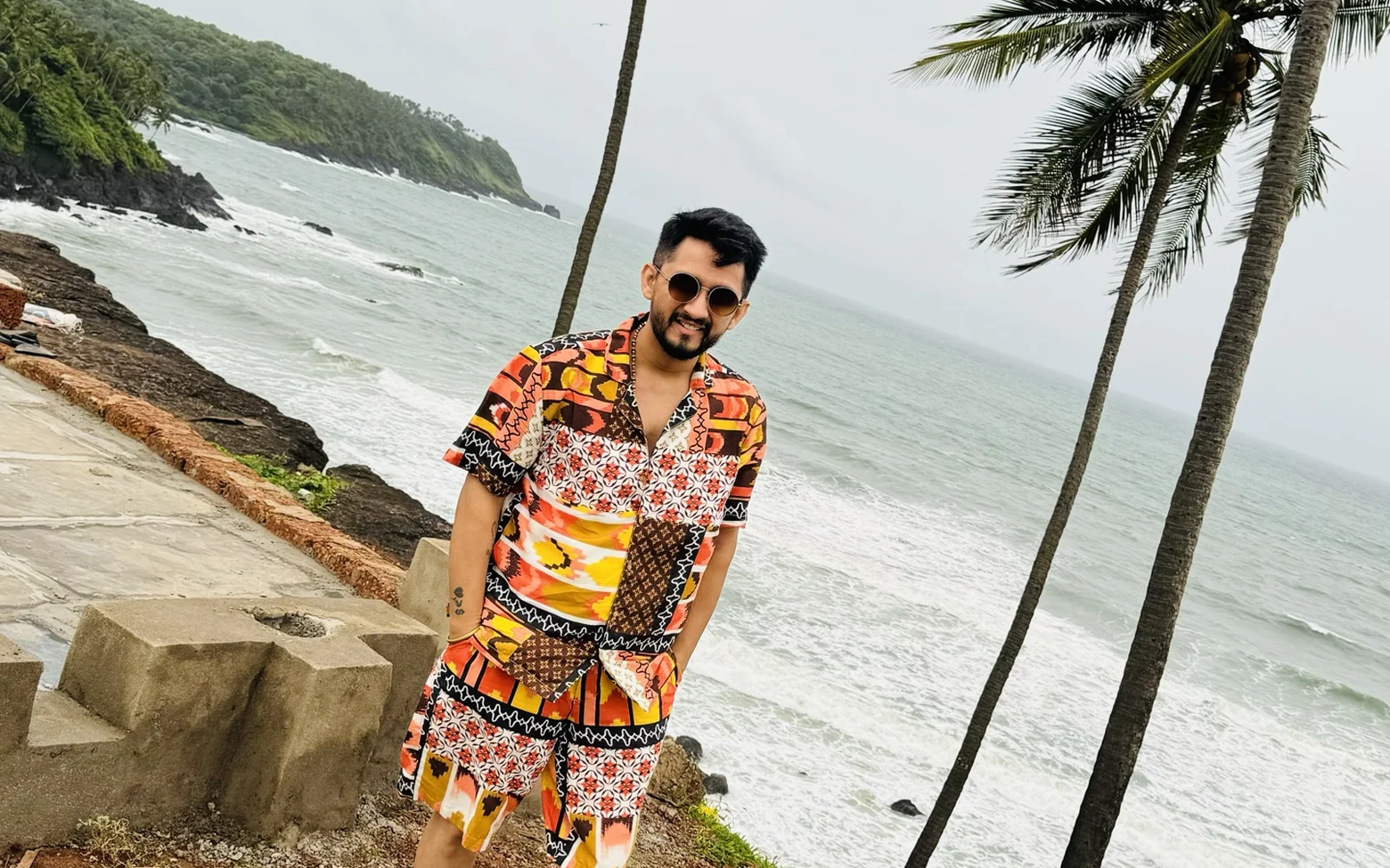 Digital Pratik smiling in sunglasses and a bright printed shirt, standing on a cliff above the sea with palm trees behind him