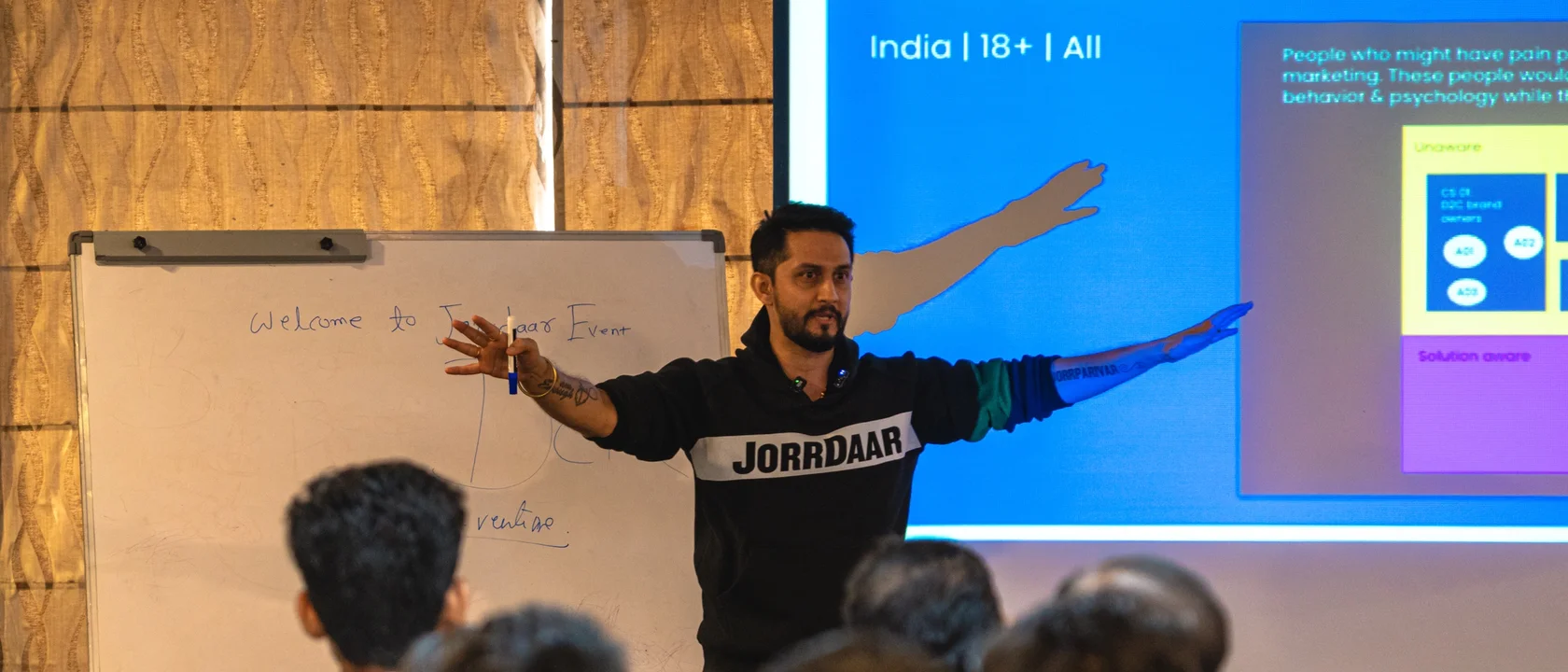 Digital Pratik teaching a room at a JorrDaar event, arms wide, a whiteboard and a slide behind him