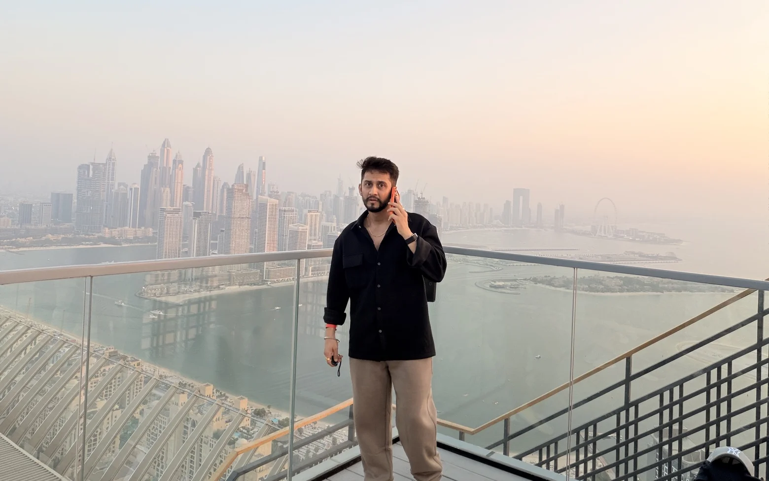 Digital Pratik on a phone call on a high rooftop deck, the Dubai skyline and the sea behind him