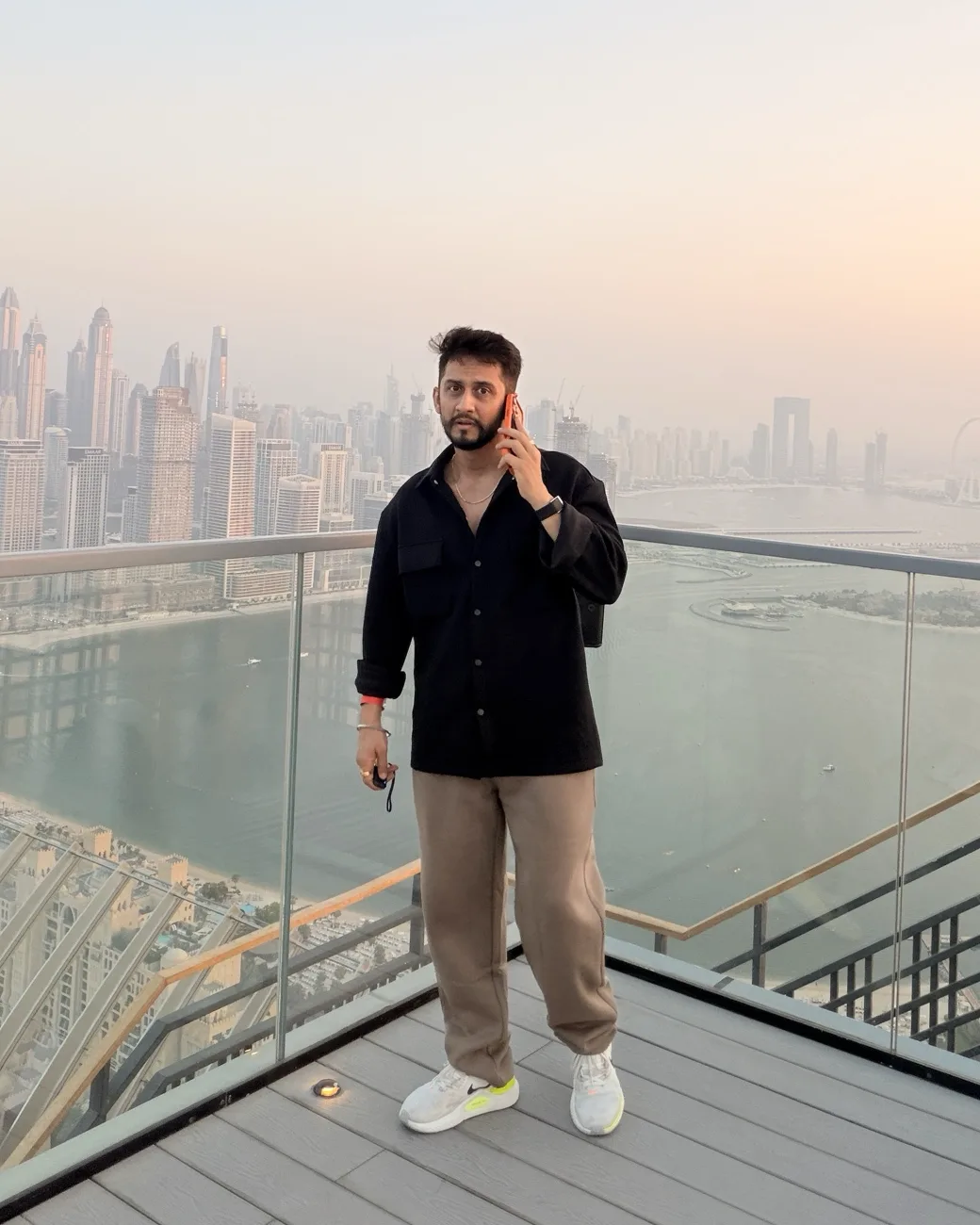 Digital Pratik on a phone call on a rooftop deck in Dubai, looking into the camera, the city behind him