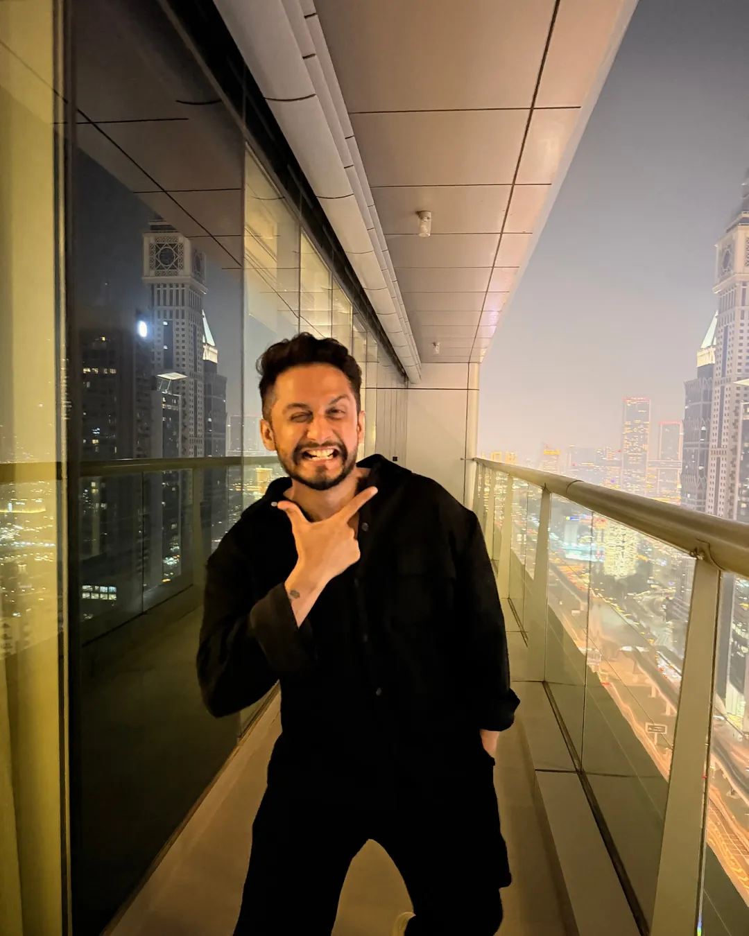 Digital Pratik smiling on a high balcony in Dubai at dusk, the towers behind him