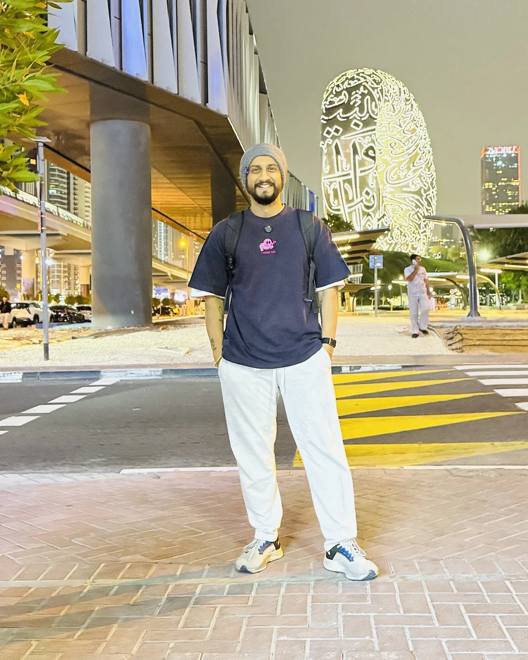 Digital Pratik on a Dubai street at night, a lit round building behind him