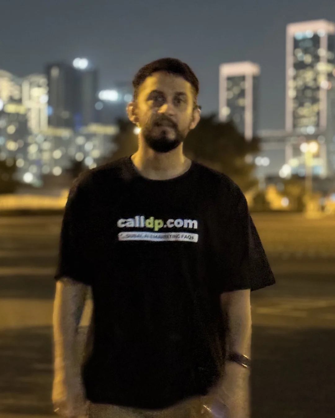 Digital Pratik at night in a calldp.com t-shirt, lit towers behind him