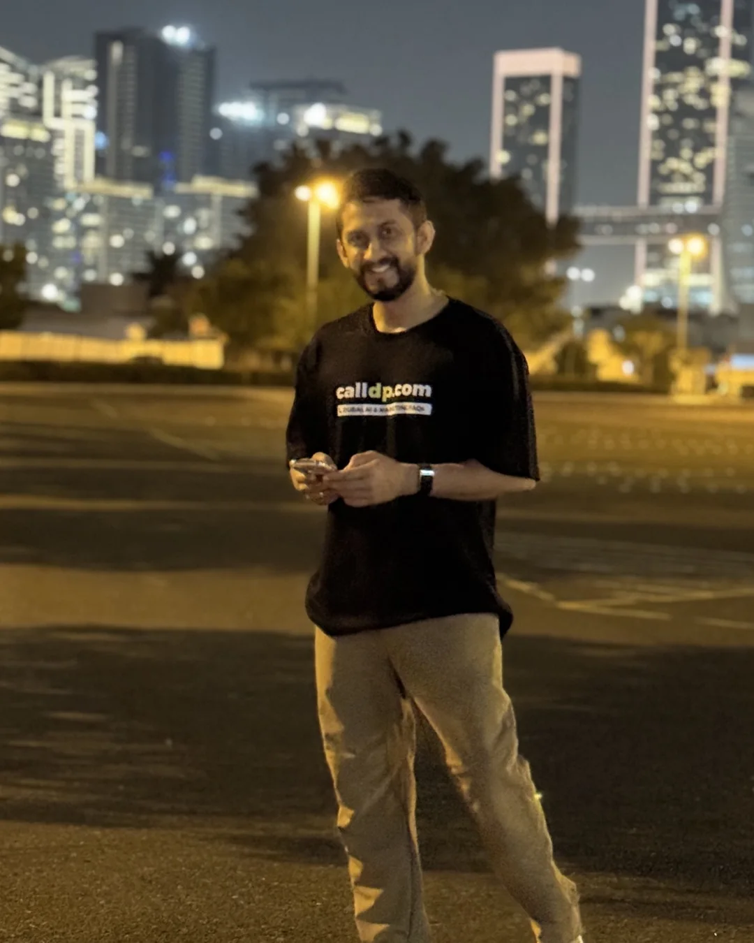 Digital Pratik smiling at night with his phone in his hand, lit towers behind him